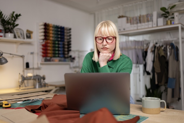 A woman sits at a table in front of an open laptop. She looks at the laptop screen with her chin resting on her fist. In the background is a white-walled room with a rack of clothes along one wall and rows of bobbins of thread in every color mounted on the other wall. The woman has shoulder-length white-blonde hair and wears a green shirt and red-rimmed glasses.